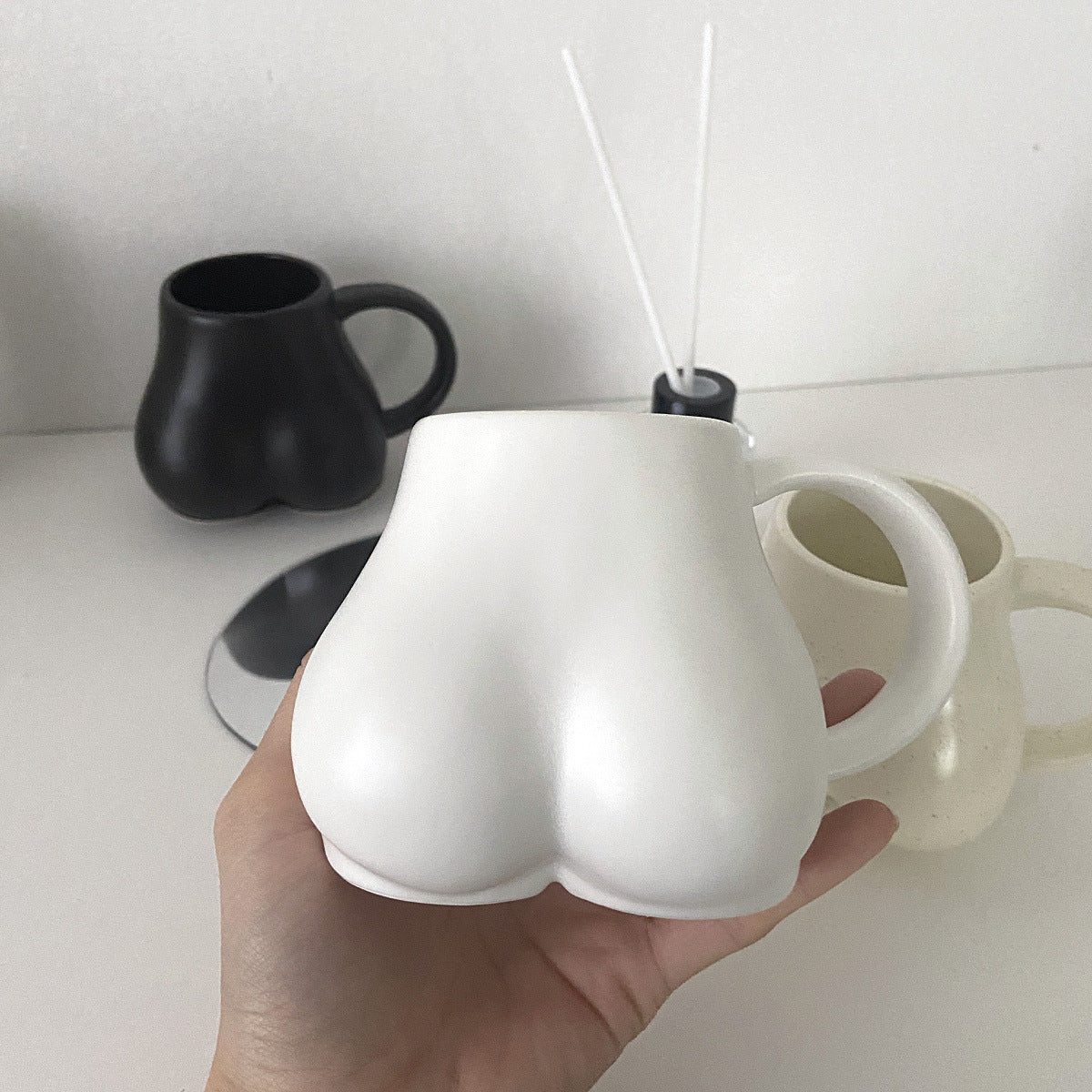 White ceramic cup shaped like a butt held by a hand on a white surface with black and beige cups in the background.